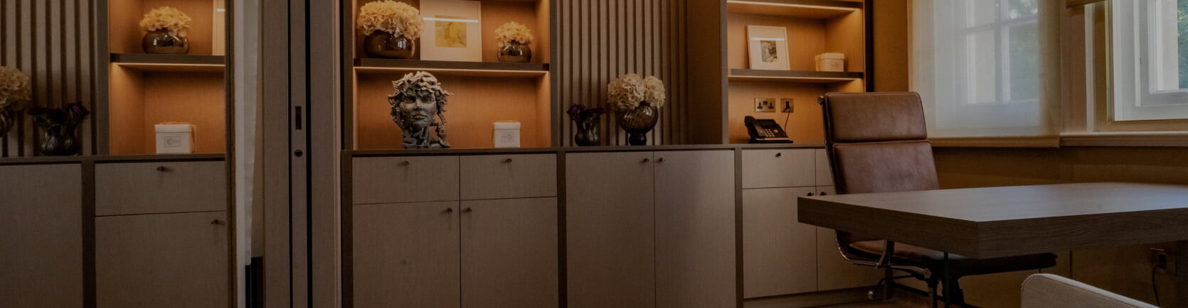 A modern, elegantly designed office space with warm ambient lighting at The London Scar Clinic, Harley Street, London. The room features built-in wooden shelves adorned with decorative items, including vases with flowers, framed pictures, and a sculpted bust. A sleek wooden desk with a brown leather chair sits near large windows with sheer curtains, allowing soft natural light to filter in.