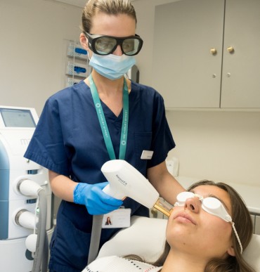 A healthcare professional in navy scrubs, protective eyewear, and a face mask performs a laser treatment on a patient's face. The patient, lying on a treatment bed with protective goggles, receives the procedure in a clinical setting.