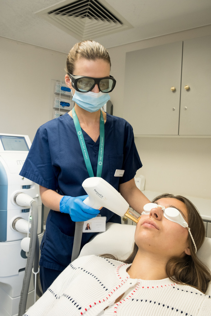 A healthcare professional in navy scrubs, protective eyewear, and a face mask performs a laser treatment on a patient's face. The patient, lying on a treatment bed with protective goggles, receives the procedure in a clinical setting.