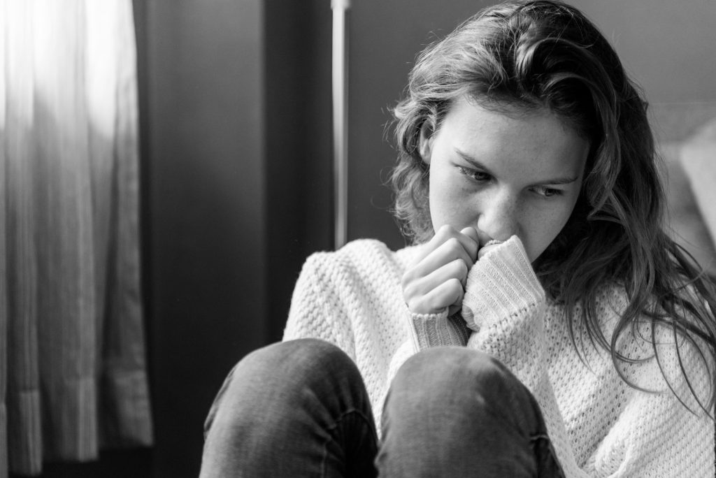 Black and white image of a young woman sitting with her knees up, looking anxious and deep in thought, symbolising emotional struggle and the journey of self-harm recovery.