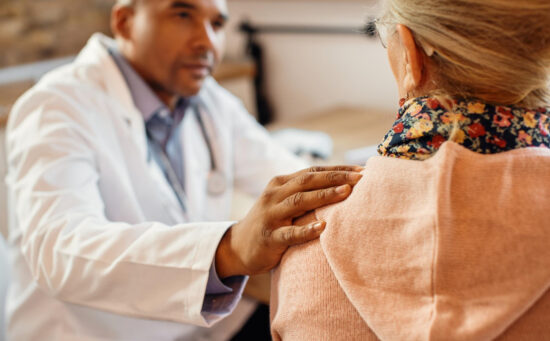 A doctor in a white coat places a comforting hand on a patient’s shoulder during a consultation.