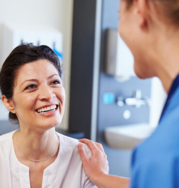 A nurse touches a patient’s shoulder while the patient smiles warmly during a consultation.